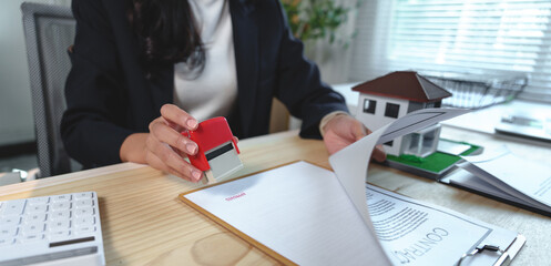 Close-up view of a female real estate agent stamping an approved mark on a contract for a new house purchase, with a miniature house model placed on the desk