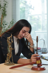 Young asian lawyer is having a bad headache, massaging her temples while sitting at her desk in the office with themis statue, scales and gavel