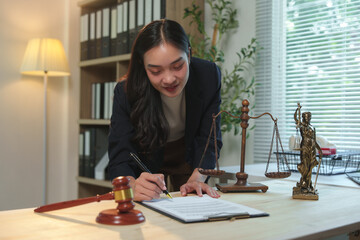 Female lawyer reviewing and signing legal document or contract in office with scales of justice and gavel on desk, ensuring fairness and legality