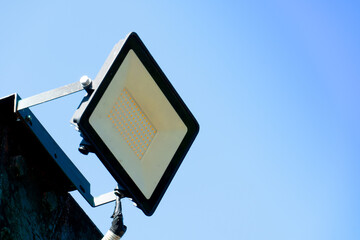 Rectangular outdoor floodlight mounted on a bracket against a clear blue sky. The light fixture is...