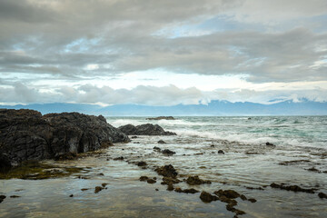 Diguisit Beach in Baler, Philippines features a rugged coastline with rocky formations dramatically positioned against the backdrop of distant mountains and a tranquil blue sea under a moody, cloud-fi