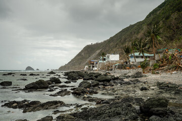 Diguisit Beach in Baler, Philippines features a rugged coastline with rocky formations dramatically positioned against the backdrop of distant mountains and a tranquil blue sea under a moody, cloud-fi