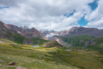 Colorful scenery with two beautiful lakes among green hills and rocks in alpine valley with view to big glacier and large snow mountain range far away under clouds in blue sky. High snowy mountains.