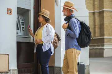 Senior couple of tourists is withdrawing money from an ATM in a bustling city setting. They appear...