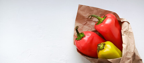 Red and green bell peppers in kraft paper are lying on a white table. Background for the advertisement of a vegetable market, farm vegetables. Space for the text.