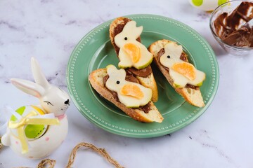 Sweet Easter sandwiches with chocolate spread and pear bunny decoration on green plate on light marble background. Easter dishes, decor.