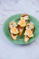 Sweet Easter sandwiches with chocolate spread and pear bunny decoration on green plate on light marble background. Easter dishes, decor.