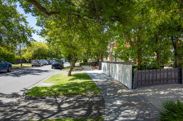A tree-lined street in a suburban neighborhood, featuring neat lawns, parked cars, and a sidewalk...