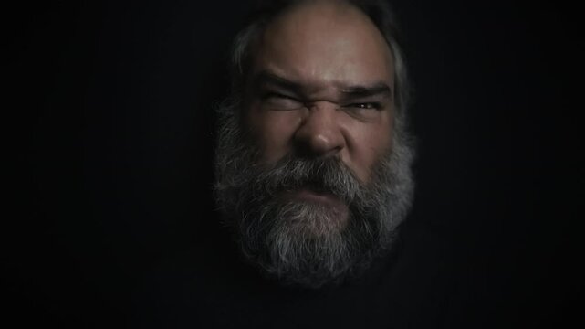 Closeup portrait of a mad, angry screaming bearded old man making faces on a black background. Sticking out tongue at camera. Fury, insanity, anger, madness topics. Slow motion video.