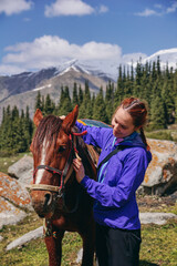 Fototapeta premium A young girl hugs a horse 's head and holds the reins. A rider and a horse in the mountains against a background of snow-capped peaks and trees
