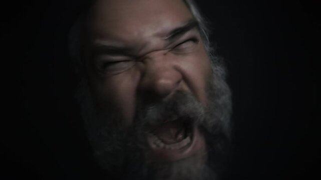 Closeup portrait of a mad, angry screaming bearded old man making faces on a black background. Fury, insanity, anger, madness topics. Slow motion video.