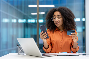 Upset and disappointed deceived woman at workplace, holding phone and bank credit card in hands,...