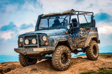 A rugged off-road vehicle covered in mud, parked on a dirt mound against a cloudy sky backdrop. © solidmaks