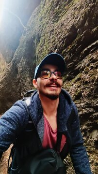 Positive man is climbing mountain between rocks and taking selfie talking to camera filmed in vertical. Smiling male tourist films vlog about trail in Georgian cliffs. Extreme leisure time activity