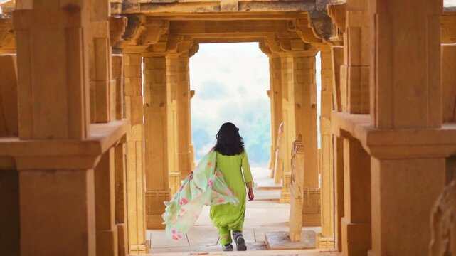 Indian young woman wearing traditional salwar suit running between pillars of Bada Bagh Cenotaphs in Jaisalmer, Rajasthan, India. Indian heritage, architecture and travel concept