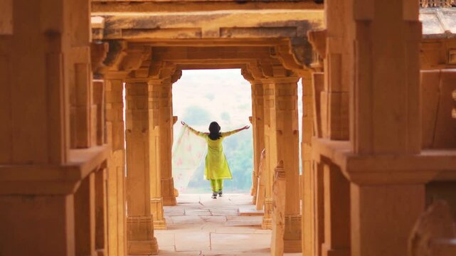 Rear view of Indian woman wearing Salwar Suit and raising arms while standing between architectural columns of Bada Bagh, Jaisalmer, India. Travel and holidays concept.