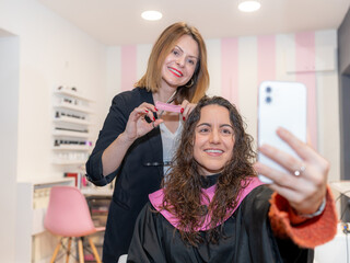 A cheerful client takes a selfie while her hairstylist trims her wet hair in a vibrant and modern...