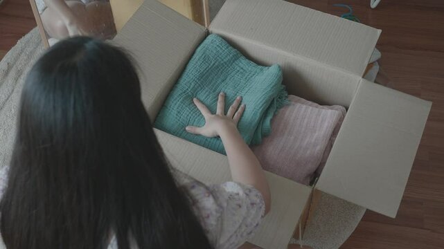 A woman is seen organizing her clothes into a cardboard box while looking in a mirror. This scene captures the essence of decluttering and preparing for a fresh start, mixed with personal reflection.