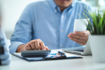 A man is sitting at a desk with a calculator and a receipt in his hand. He is focused on the...
