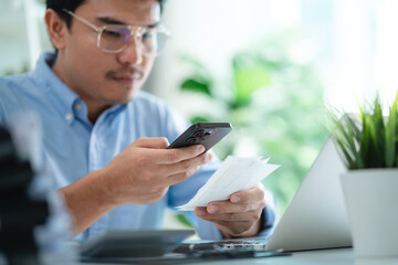 A man is sitting at a desk with a laptop and a cell phone. He is looking at the cell phone and he is checking something on it