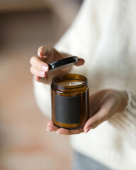Aroma soy candle in brown glass jar with black lid in woman's hands. Amber minimalist candle packaging design with copy space. Aromatherapy, craft home decor, gift idea, home comfort, relaxation.