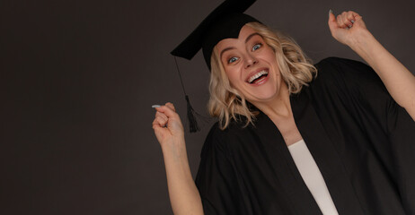 A cheerful female graduate in a black gown and cap with tassel celebrates her achievement, smiling...