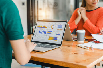 laptop displaying colorful data charts and graphs is placed on wooden desk, ed by office supplies and coffee cups, with two people collaborating in modern office space