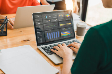 person working on laptop displaying data analytics dashboards with charts and graphs, sitting at wooden desk with documents, coffee cup, and another person using laptop in background