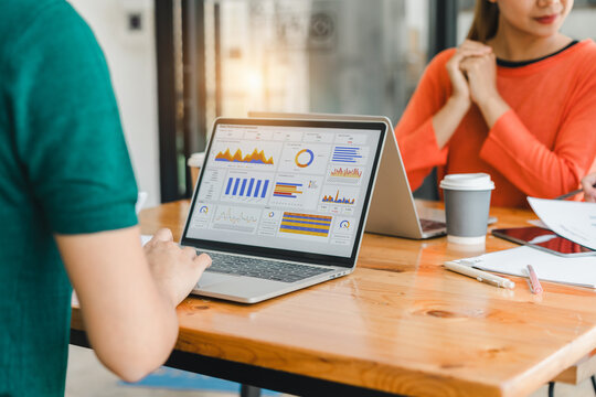 laptop displaying colorful data charts and graphs is placed on wooden desk, ed by office supplies and coffee cups, with two people collaborating in modern office space