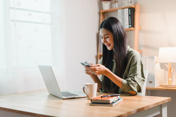 young woman smiling while using her smartphone at wooden desk with laptop, coffee mug, and books in bright home office