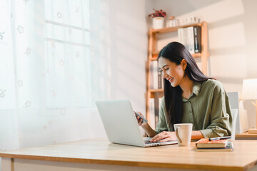 woman working on laptop at wooden desk in bright home office, smiling while holding smartphone,...