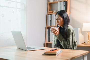 young woman sits at wooden desk, holding cup while looking at laptop screen in cozy home office with bookshelves and soft lighting