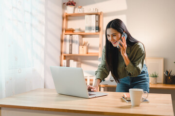 cheerful woman multitasking while talking on phone and working on laptop at wooden desk in bright home office with shelves and decor in background