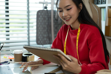 A woman in a red sweater is sitting at a desk with a laptop and a notebook. She is smiling and she...