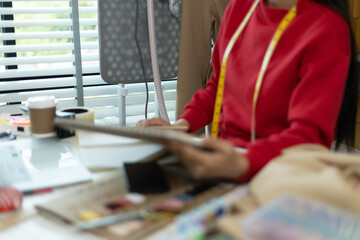 A woman in a red sweater is sitting at a desk with a laptop and a notebook. She is smiling and she is working on something. The desk is cluttered with various items such as a cup, a cell phone
