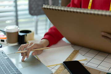 A woman in a red sweater is sitting at a desk with a laptop and a notebook. She is smiling and she...