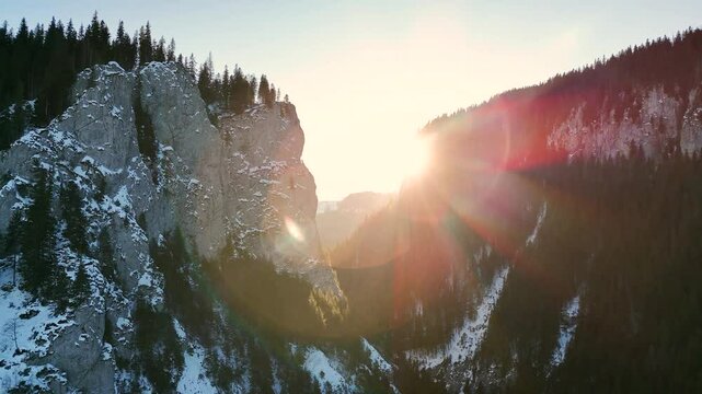 Aerial drone footage of snow-covered cliffs and pine forests at sunset in Bucegi Natural Park, Romania, showcasing a dramatic winter mountain landscape