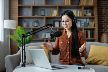 Asian woman hosting a podcast at home, speaking into a microphone while using a laptop. Wearing...