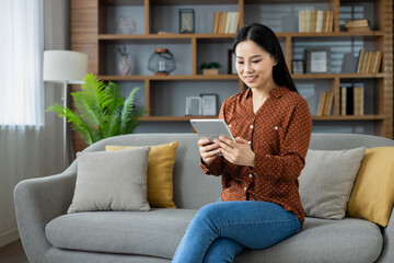 Young Asian woman sitting at home on the couch smiling happily using a tablet computer, using an educational app and reading online books, watching video streams.