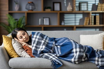 Obraz premium Asian woman resting peacefully on couch, wrapped in cozy blanket, evoking sense of relaxation and comfort in home environment. Perfect depiction of tranquility and restful living.