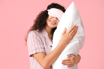 Beautiful young African-American woman in pajamas and mask with soft pillow on pink background....