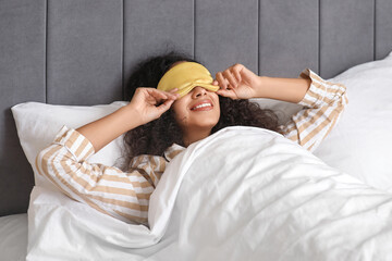 Beautiful young African-American woman in pajamas and mask lying on bed in bedroom. World Sleep Day