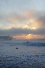 Naklejka premium Winter sunset, through the mist and over the smoking ocean. A surfer goes into the water. Anglet, France