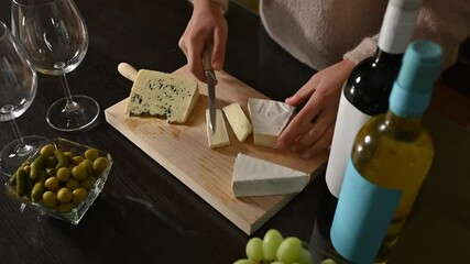 Woman slices cheese on a wooden board. She is preparing a cheese and wine appetizer platter with grapes, olives and pickles.