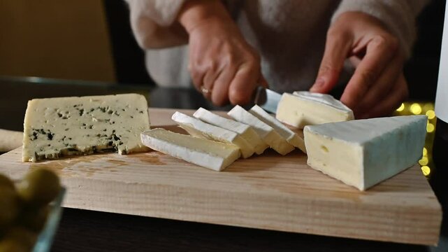 Woman slices brie cheese on a wooden cutting board. Close up shot of hands cutting cheese with soft lighting. Olives and blue cheese are visible in the background.