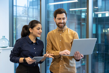 Two colleagues man and woman inside office standing by window talking, group of office workers...