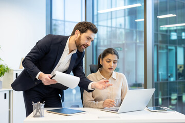 Mentor man in business suit helping colleague female intern at workplace, employees members of business team serious communicating at workplace discussing project, business meeting.
