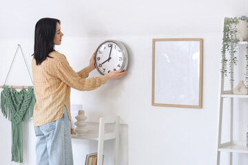 Young woman hanging clock on light wall at home
