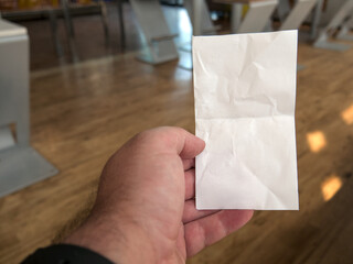 Close-up shot of a male hand holding a slightly crumpled white paper bill in a restaurant, personal perspective. Copy space, template, mockup