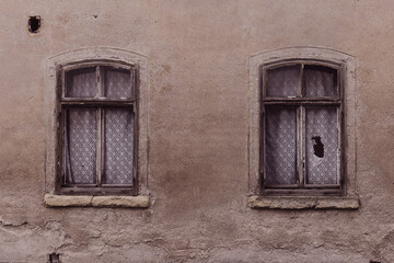 Two worn wooden windows with lace curtains on a weathered wall in an old building